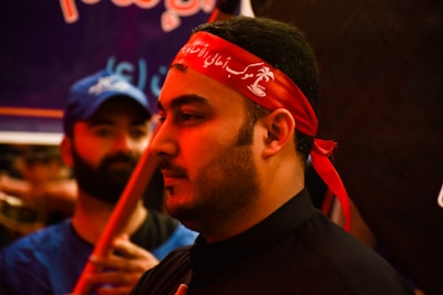 The image shows a close-up of a man wearing a red headband with white script. His expression is calm and focused. In the background, another person in a blue shirt and cap is visible, slightly out of focus.