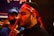 The image shows a close-up of a man wearing a red headband with white script. His expression is calm and focused. In the background, another person in a blue shirt and cap is visible, slightly out of focus.