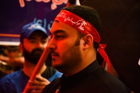 The image shows a close-up of a man wearing a red headband with white script. His expression is calm and focused. In the background, another person in a blue shirt and cap is visible, slightly out of focus.