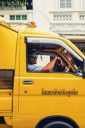 A person is seated inside a bright yellow truck with black text in Thai on the side. They are wearing a cap and sunglasses, and appear to be smiling. The truck is parked in front of a building with decorative elements and windows. There are some plants visible on a ledge.