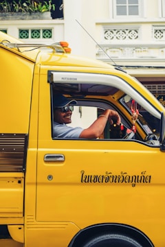 A person is seated inside a bright yellow truck with black text in Thai on the side. They are wearing a cap and sunglasses, and appear to be smiling. The truck is parked in front of a building with decorative elements and windows. There are some plants visible on a ledge.