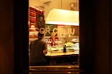 A warm, dimly lit restaurant scene viewed through a window, featuring two people sitting at a bar counter. Several glasses of wine and plates of food are on display, while shelves stocked with bottles line the background. The overall ambiance is cozy and intimate, enhanced by the soft lighting.