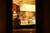 A warm, dimly lit restaurant scene viewed through a window, featuring two people sitting at a bar counter. Several glasses of wine and plates of food are on display, while shelves stocked with bottles line the background. The overall ambiance is cozy and intimate, enhanced by the soft lighting.