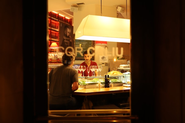 A warm, dimly lit restaurant scene viewed through a window, featuring two people sitting at a bar counter. Several glasses of wine and plates of food are on display, while shelves stocked with bottles line the background. The overall ambiance is cozy and intimate, enhanced by the soft lighting.
