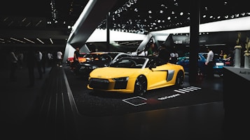 A sleek yellow sports car is prominently displayed on a showroom floor surrounded by other vehicles and people. The environment is modern and well-lit, indicating an automotive exhibition.