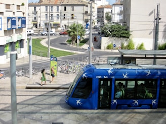 A blue tram with white bird designs is moving through an urban area with people inside. Two women are walking on the pavement near a bicycle rental station. Several bicycles are parked, and the street is lined with greenery and buildings in the background.