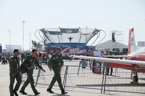 Four people in military flight suits walk past a fenced area on what appears to be an airfield. In the background, a trampoline or similar device is seen with a person in mid-jump. There are crowds of people, possibly at an air show or military display event. A small aircraft with visible markings is parked nearby, and various tents and banners display advertisements.