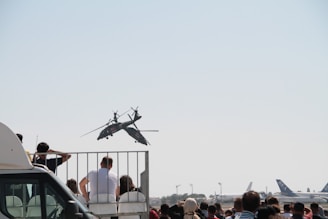 Crowd gathered to watch an aerobatic display by a helicopter performing stunts in a clear sky. The scene includes people standing on a platform and more individuals on the ground, with commercial aircraft parked in the distance.