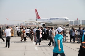 A large group of people is gathered on a tarmac near a Turkish Cargo airplane. The aircraft is prominently displayed in the background, parked close to the barriers where spectators stand. Many individuals are taking pictures or observing closely. Tall buildings can be seen in the distant background, indicating the setting is near an urban area.