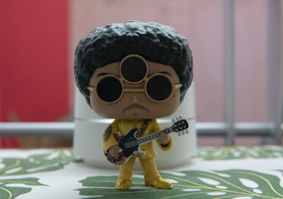 A stylized figurine with a large afro hairstyle and sunglasses, holding a guitar. It is dressed in a glittery gold outfit, standing on a surface with a leafy pattern. The background features blurred elements in red and gray with a metallic railing.