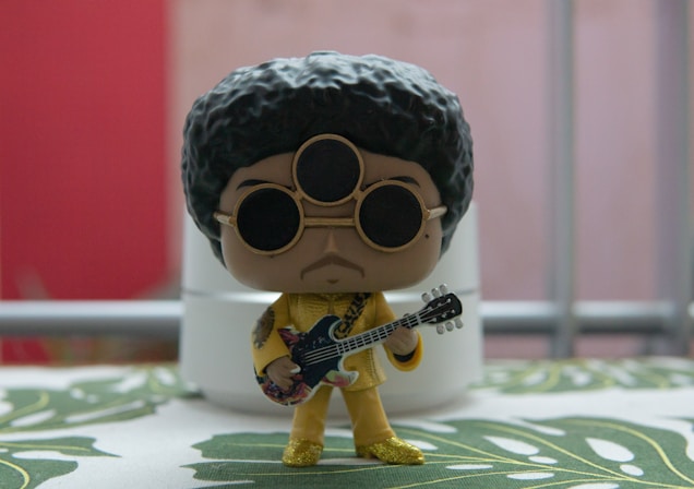 A stylized figurine with a large afro hairstyle and sunglasses, holding a guitar. It is dressed in a glittery gold outfit, standing on a surface with a leafy pattern. The background features blurred elements in red and gray with a metallic railing.