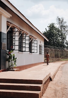 A person dressed in a brown uniform is walking along a path next to a building with large windows and security bars. The building has a white exterior with some greenery at the base of the wall. There is a wire fence and trees in the background under a partly cloudy sky.