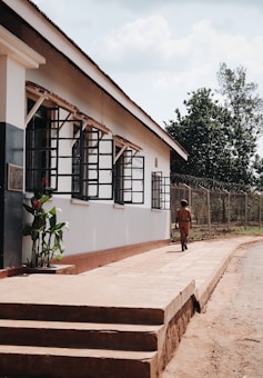 A person dressed in a brown uniform is walking along a path next to a building with large windows and security bars. The building has a white exterior with some greenery at the base of the wall. There is a wire fence and trees in the background under a partly cloudy sky.