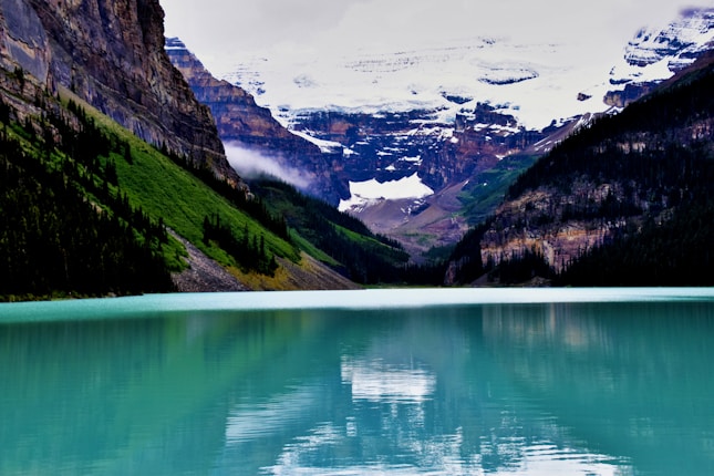 A pristine turquoise lake is surrounded by steep, forested mountains. In the distance, snow-capped peaks rise majestically under a cloudy sky, creating a serene and picturesque natural landscape.