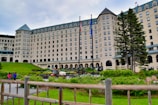 A grand, multi-story hotel with a castle-like architectural style surrounded by well-kept gardens and green lawns. Tall trees stand on the right side, and flags are prominently displayed in front of the building. A few people are visible alongside a vehicle that appears to be for garden maintenance.