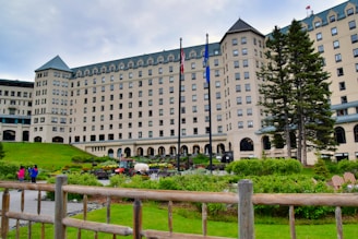 A grand, multi-story hotel with a castle-like architectural style surrounded by well-kept gardens and green lawns. Tall trees stand on the right side, and flags are prominently displayed in front of the building. A few people are visible alongside a vehicle that appears to be for garden maintenance.