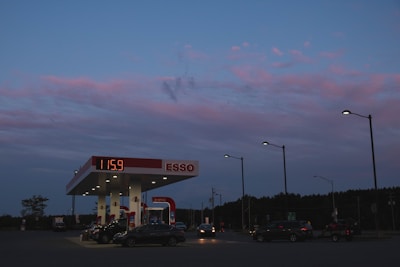 A gas station is illuminated under a twilight sky with hues of blue and pink. Several cars are parked under the gas station canopy, with lit streetlights lining the area. The price sign displays the fuel cost, and trees can be seen in the background.