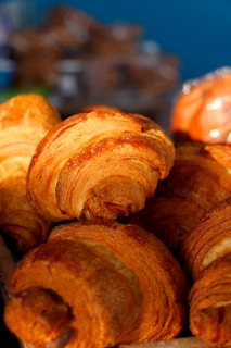 Freshly baked croissants with a golden-brown crust are displayed in a basket. The pastries exhibit a flaky and layered texture, suggesting they are well-cooked and appetizing. The background is blurred, focusing attention on the croissants.