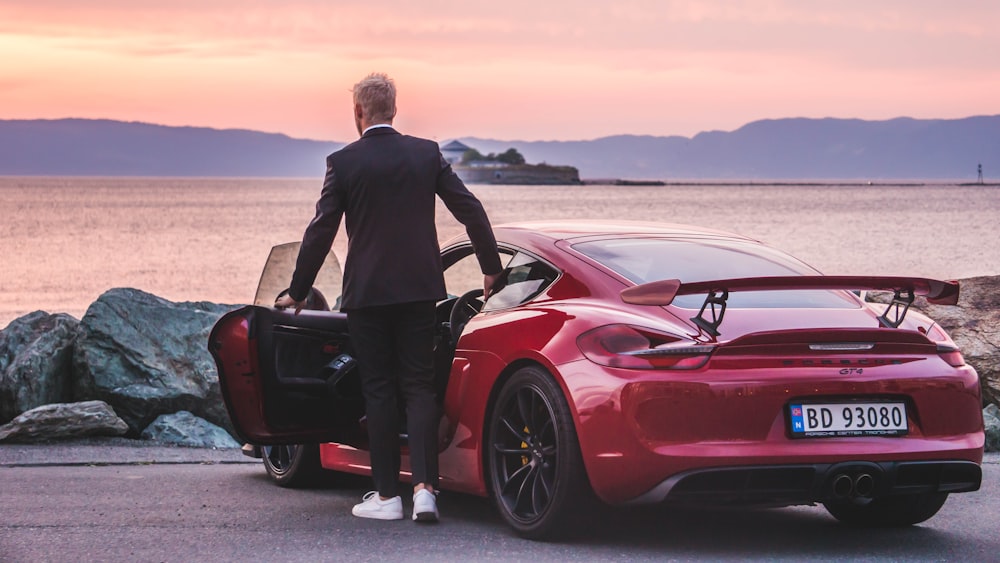 man standing beside a red coupe by the sea