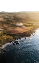 A coastal landscape during sunset featuring a vast expanse of grassy fields and undulating hills, with a winding road leading to a solitary house near the edge of the land. The rocky shoreline meets dark blue ocean waters, creating a serene and tranquil scene.