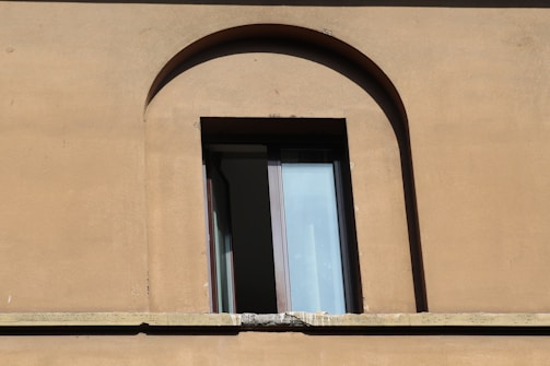 A single rectangular window with a partially open glass pane set in a brown, stucco wall. The window frame is dark and it is situated in an arched alcove of the building's facade. The wall has some visible texture and weathering.