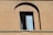 A single rectangular window with a partially open glass pane set in a brown, stucco wall. The window frame is dark and it is situated in an arched alcove of the building's facade. The wall has some visible texture and weathering.