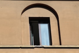 A single rectangular window with a partially open glass pane set in a brown, stucco wall. The window frame is dark and it is situated in an arched alcove of the building's facade. The wall has some visible texture and weathering.