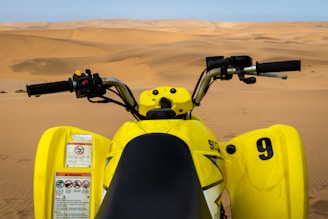 A yellow quad bike viewed from the rider's perspective, positioned on vast, undulating sand dunes. The handlebars and controls are prominently visible in the foreground, while the expansive desert landscape stretches out toward the horizon.