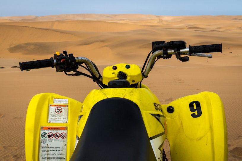 A yellow quad bike viewed from the rider's perspective, positioned on vast, undulating sand dunes. The handlebars and controls are prominently visible in the foreground, while the expansive desert landscape stretches out toward the horizon.