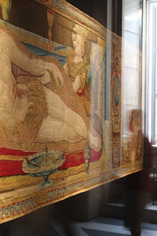 An ornate tapestry with intricate designs and vivid colors, depicting a reclining figure. The tapestry includes elements like a chalice, a winged figure, and decorative border patterns. The scene appears historical or mythological, evocative of classical art styles. Reflections of the surroundings are visible, suggesting the tapestry is displayed in a setting with glass or other reflective surfaces.