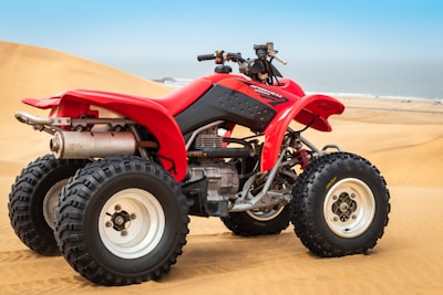 A red all-terrain vehicle (ATV) with large, rugged tires is positioned on sandy desert dunes. The clear sky and distant view of the ocean or sea create a backdrop of open, natural space.
