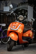 A vibrant orange scooter is parked on a cobblestone street. The scooter is adorned with yellow and white flower decals, giving it a playful and cheerful appearance. It is positioned in front of a shop window decorated with spherical ornaments and star shapes. A sign visible on the window offers rental options, including prices per day for bicycles.