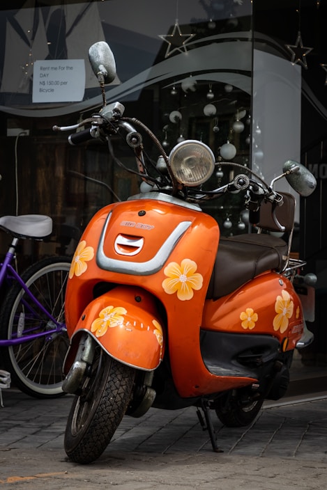 A vibrant orange scooter is parked on a cobblestone street. The scooter is adorned with yellow and white flower decals, giving it a playful and cheerful appearance. It is positioned in front of a shop window decorated with spherical ornaments and star shapes. A sign visible on the window offers rental options, including prices per day for bicycles.