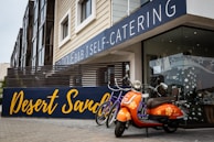 An orange scooter with floral designs is parked in front of a modern building that features a large sign reading 'Desert Sand'. There are also two bicycles nearby. The building has a contemporary design with large windows, beige siding, and a balcony with horizontal wooden slats.