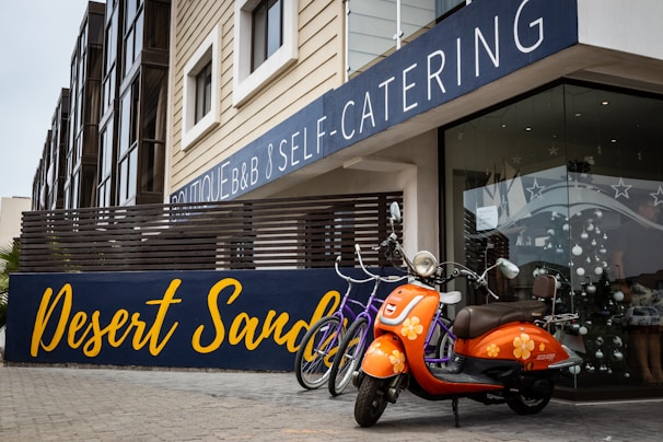 An orange scooter with floral designs is parked in front of a modern building that features a large sign reading 'Desert Sand'. There are also two bicycles nearby. The building has a contemporary design with large windows, beige siding, and a balcony with horizontal wooden slats.