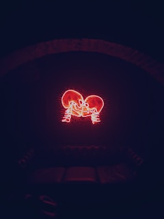 A neon art piece depicting two skeletons facing each other, positioned on a background of a dark wall. The glowing red outlines of the skeletons create an intense visual effect, with the surrounding area mostly obscured in shadows. Below the neon art, a dimly lit couch is visible, blending into the setting.