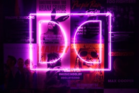 A collection of posters for various music concerts, illuminated by bright purple neon lights in the shape of the letters D and C. The posters feature artists like Ariana Grande, Imagine Dragons, Stevie Wonder, and others. The neon lights create a vibrant and electrifying atmosphere.