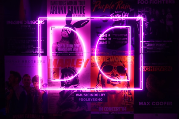 A collection of posters for various music concerts, illuminated by bright purple neon lights in the shape of the letters D and C. The posters feature artists like Ariana Grande, Imagine Dragons, Stevie Wonder, and others. The neon lights create a vibrant and electrifying atmosphere.