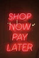 Bright pink neon sign with the words 'SHOP NOW PAY LATER' against a dark background. The sign casts a warm glow, creating a vibrant and enticing atmosphere.