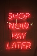 Bright pink neon sign with the words 'SHOP NOW PAY LATER' against a dark background. The sign casts a warm glow, creating a vibrant and enticing atmosphere.