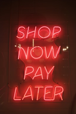Bright pink neon sign with the words 'SHOP NOW PAY LATER' against a dark background. The sign casts a warm glow, creating a vibrant and enticing atmosphere.