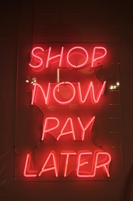 Bright pink neon sign with the words 'SHOP NOW PAY LATER' against a dark background. The sign casts a warm glow, creating a vibrant and enticing atmosphere.