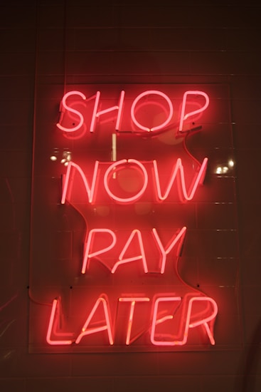 Bright pink neon sign with the words 'SHOP NOW PAY LATER' against a dark background. The sign casts a warm glow, creating a vibrant and enticing atmosphere.