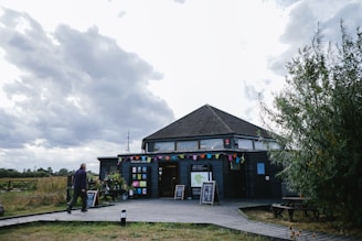 A wooden visitor center with a hexagonal roof sits under a cloudy sky. The building is decorated with colorful triangular pennant banners. There are various signs and placards in front, along with some potted plants. A man is walking towards the visitor center on a wooden deck that leads up to it. Surrounding the area is grassy terrain and some bushes.