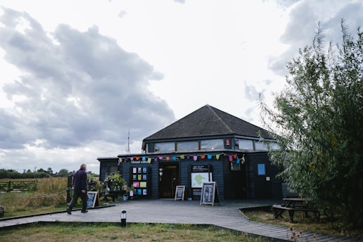 A wooden visitor center with a hexagonal roof sits under a cloudy sky. The building is decorated with colorful triangular pennant banners. There are various signs and placards in front, along with some potted plants. A man is walking towards the visitor center on a wooden deck that leads up to it. Surrounding the area is grassy terrain and some bushes.