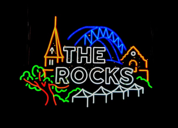 Neon sign featuring the text 'The Rocks' with colorful icons including a church spire, a bridge arch, a building, a tree, and a set of tents or canopies outlined in neon lights against a black background.