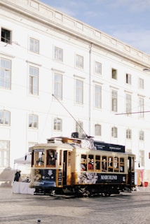 A vintage tram, adorned with advertising, travels along a cobblestone street in front of a white, multi-story historic building with numerous windows. People are visible inside the tram, and a few market stalls are set up nearby.