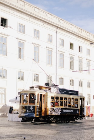 A vintage tram, adorned with advertising, travels along a cobblestone street in front of a white, multi-story historic building with numerous windows. People are visible inside the tram, and a few market stalls are set up nearby.
