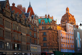 A row of ornate, historic buildings with intricate architectural details is visible, featuring large windows and decorative facades. One building is prominently marked with the name 'Harrods' in large letters. Another building has a distinctive dome and is draped in flags. A large advertisement for a Samsung Galaxy phone is displayed on the side of a building. Street lights line the road in front of these buildings.