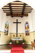 An indoor setting with a central altar decorated with white flowers and candles. Two colorful stained glass windows flank the altar, depicting religious scenes. A large black cross is mounted on the wall above the altar. The ceiling features dark wooden beams, and a red carpet leads up to the altar.